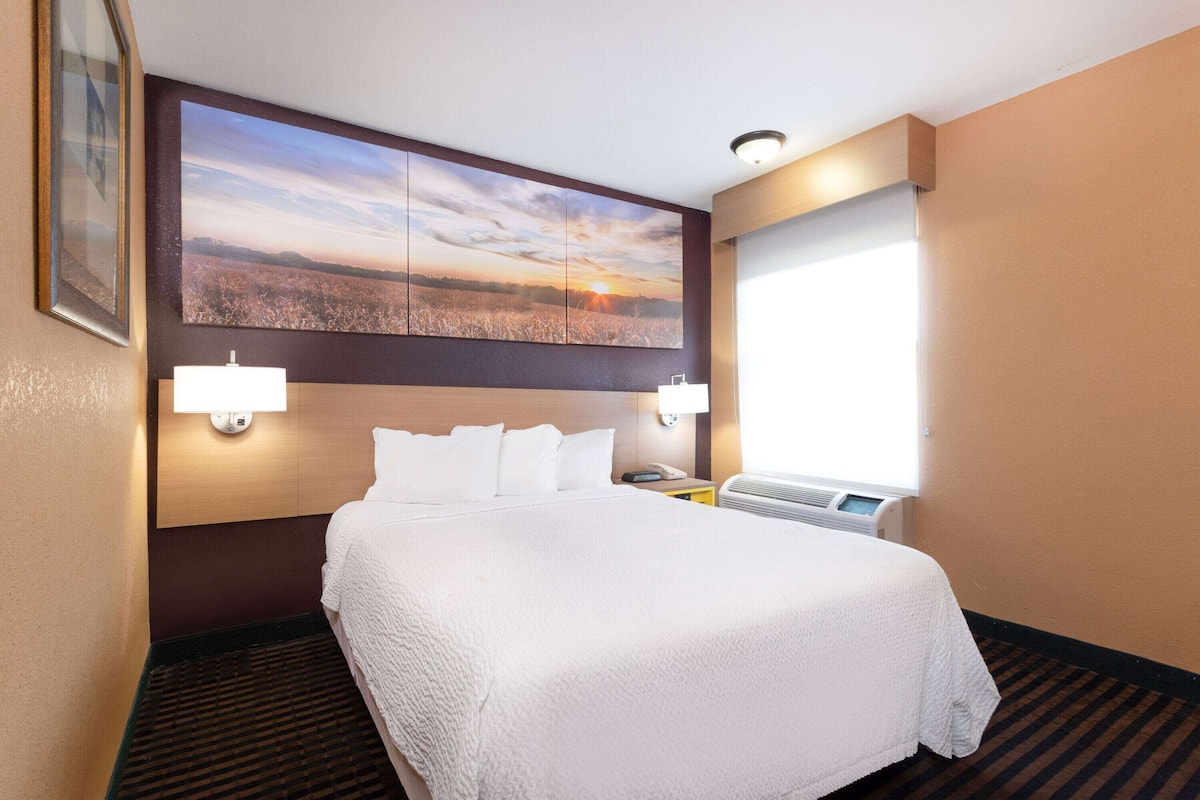 Habitación de hotel con una cama grande, dos lámparas de noche, una mesita de noche, una ventana con persianas y un cuadro colgado en la pared que muestra un atardecer sobre un campo.