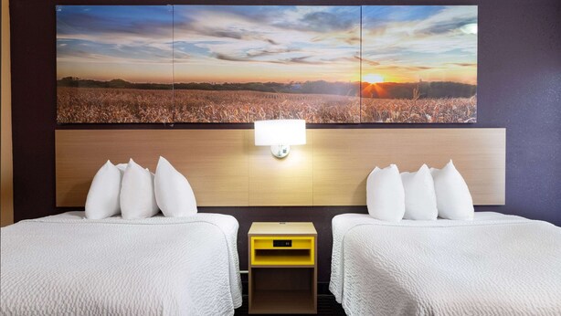 A hotel room with two beds, a nightstand, a wall-mounted lamp, and a large landscape painting. - Days Inn by Wyndham Anderson IN (Anderson)