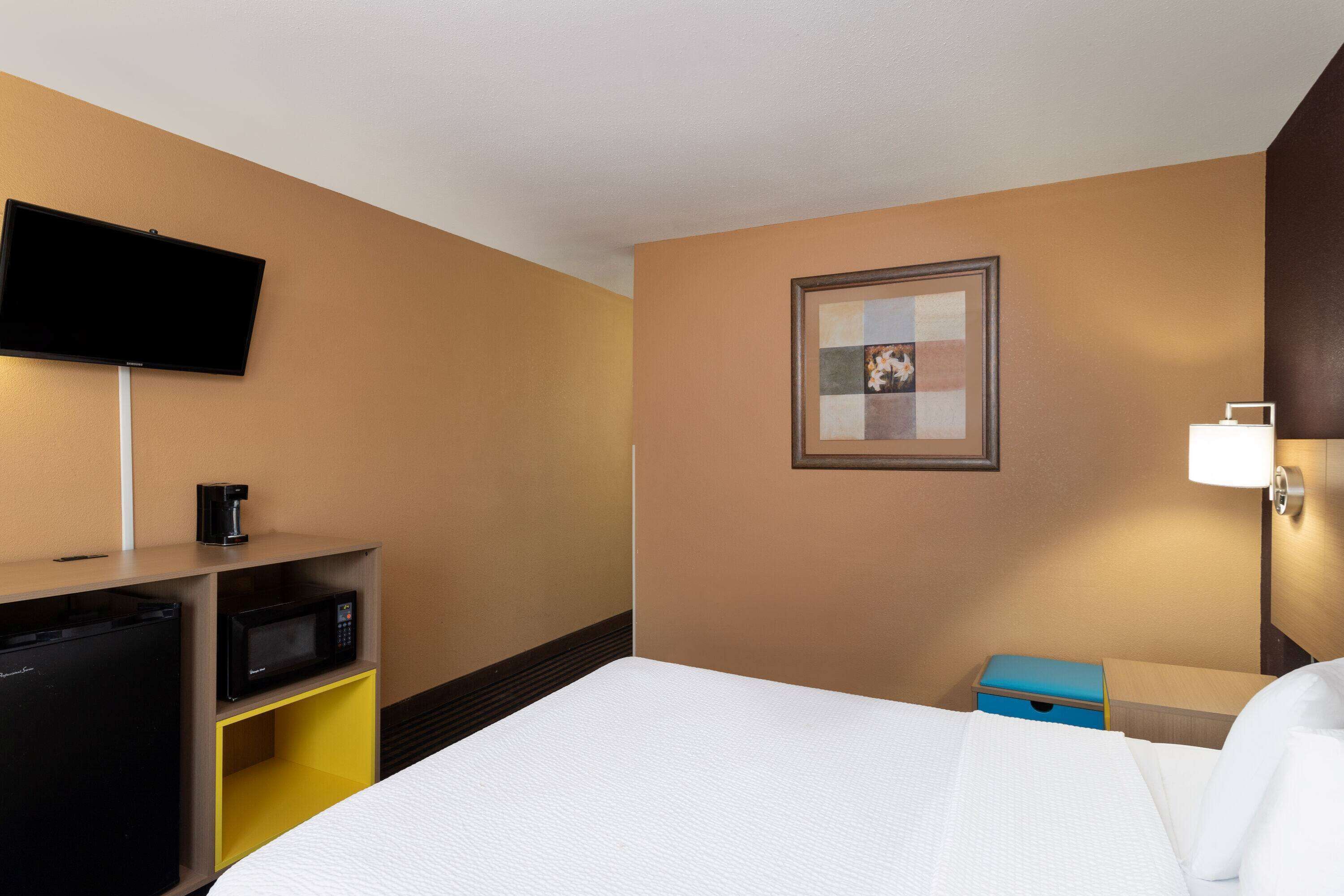 Habitación de hotel con cama, un televisor de pantalla plana en la pared, microondas, cafetera y un pequeño caja azul para guardar objetos.