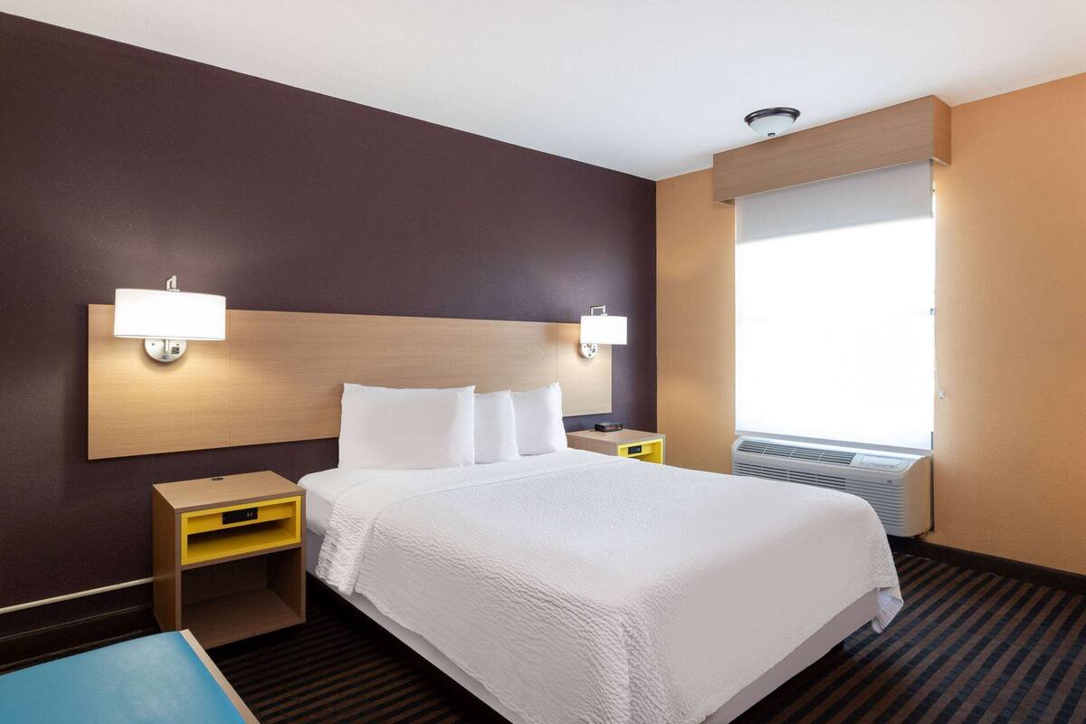 Habitación de hotel con una cama grande, dos mesitas de noche, un aparador, una ventana con persianas y un aire acondicionado instalado en la pared.