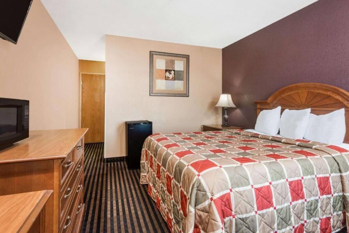 Una habitación de hotel con una cama, televisión, cómoda, cubo de basura y un cuadro en la pared.