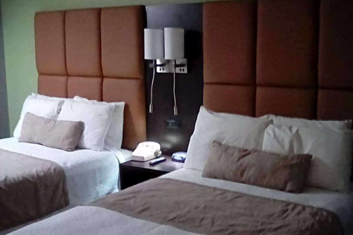 A hotel room with two beds, each with white linens and beige pillows. There is a nightstand with a telephone and a lamp between the beds.