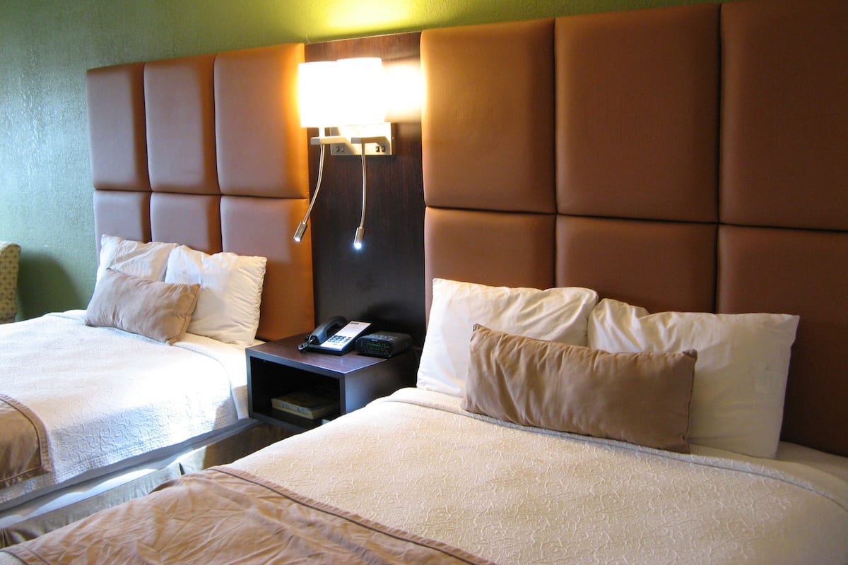 A hotel room with two beds, a nightstand with a phone, and a wall-mounted lamp.