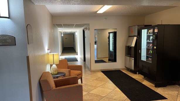 A hotel lobby with a reception desk, a seating area with a chair and a lamp, a refrigerator, and a vending machine. - Nichols Inn of Hastings (Hastings)
