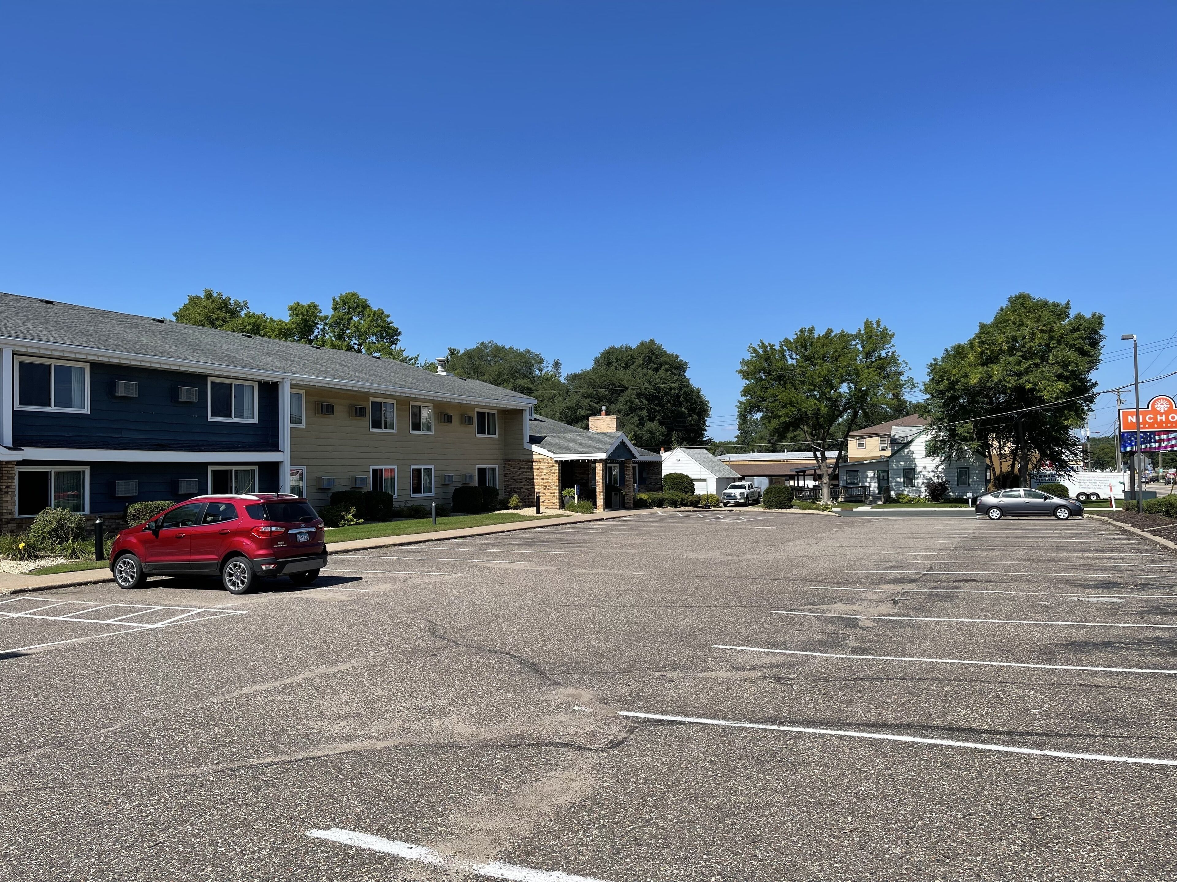 Un parking avec un SUV rouge, un petit bâtiment sur lequel on peut voir une enseigne, et une rangée de maisons.