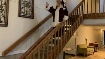 Una persona disfrazada de Papá Noel baja por una escalera de madera con barandilla, que conduce a un rellano del segundo piso con una pequeña mesa y sillas.