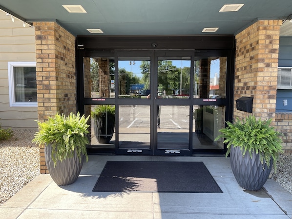 A building entrance with a glass door, two potted plants, and a black mat.
