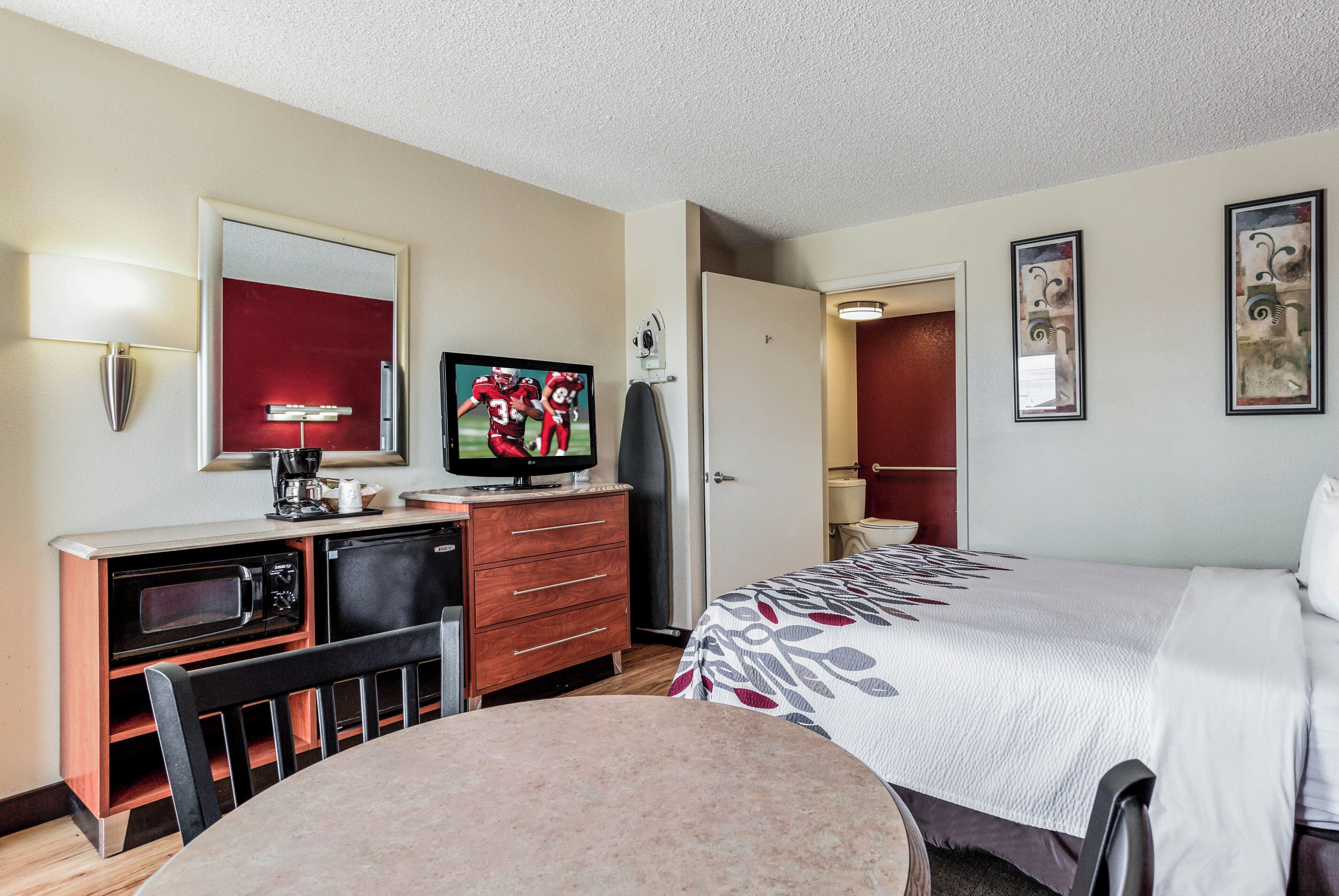 A hotel room with a bed, a desk with a television, a chair, a small table, and a door leading to the bathroom.
