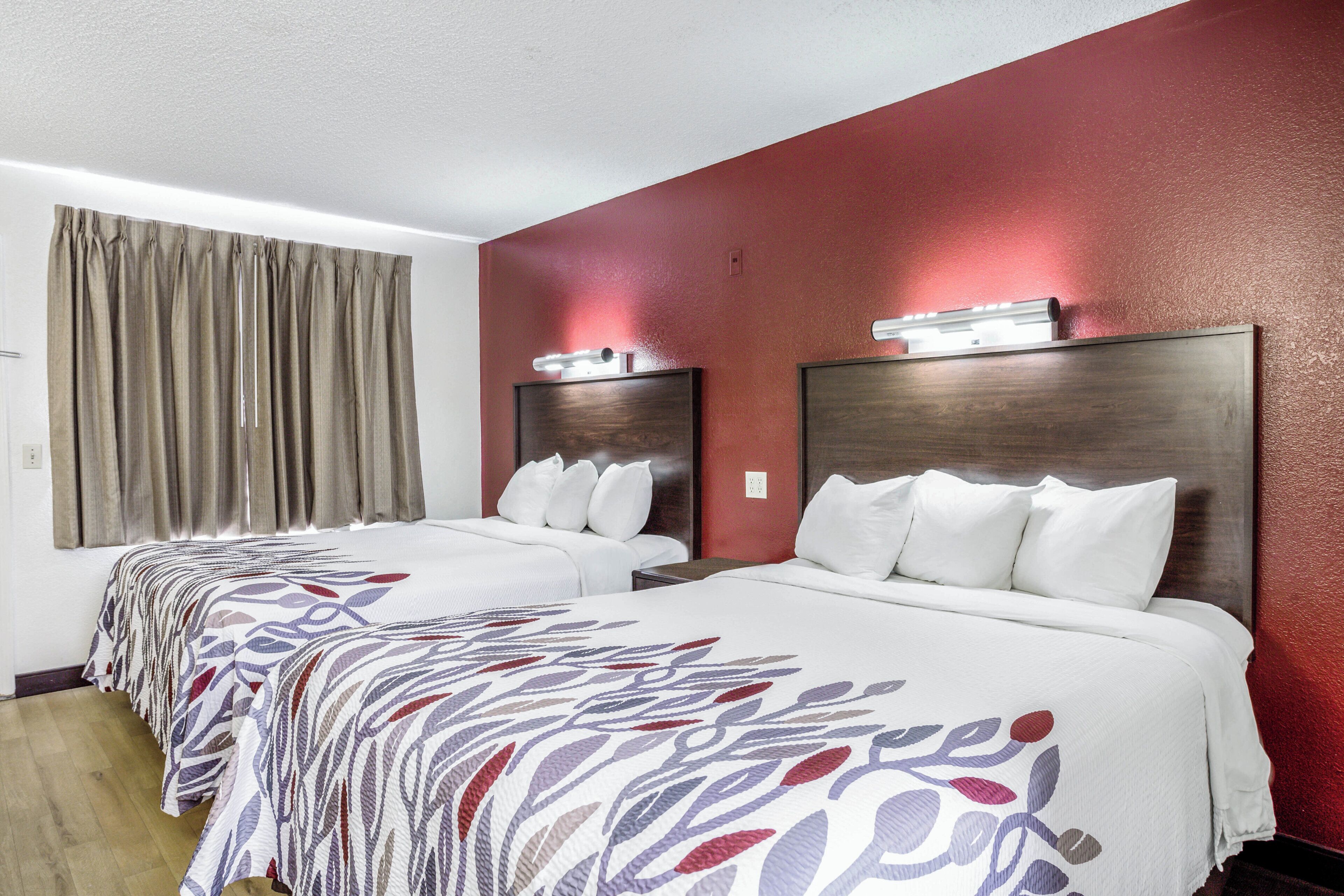 A hotel room with two beds, a large window with curtains, and a red accent wall.