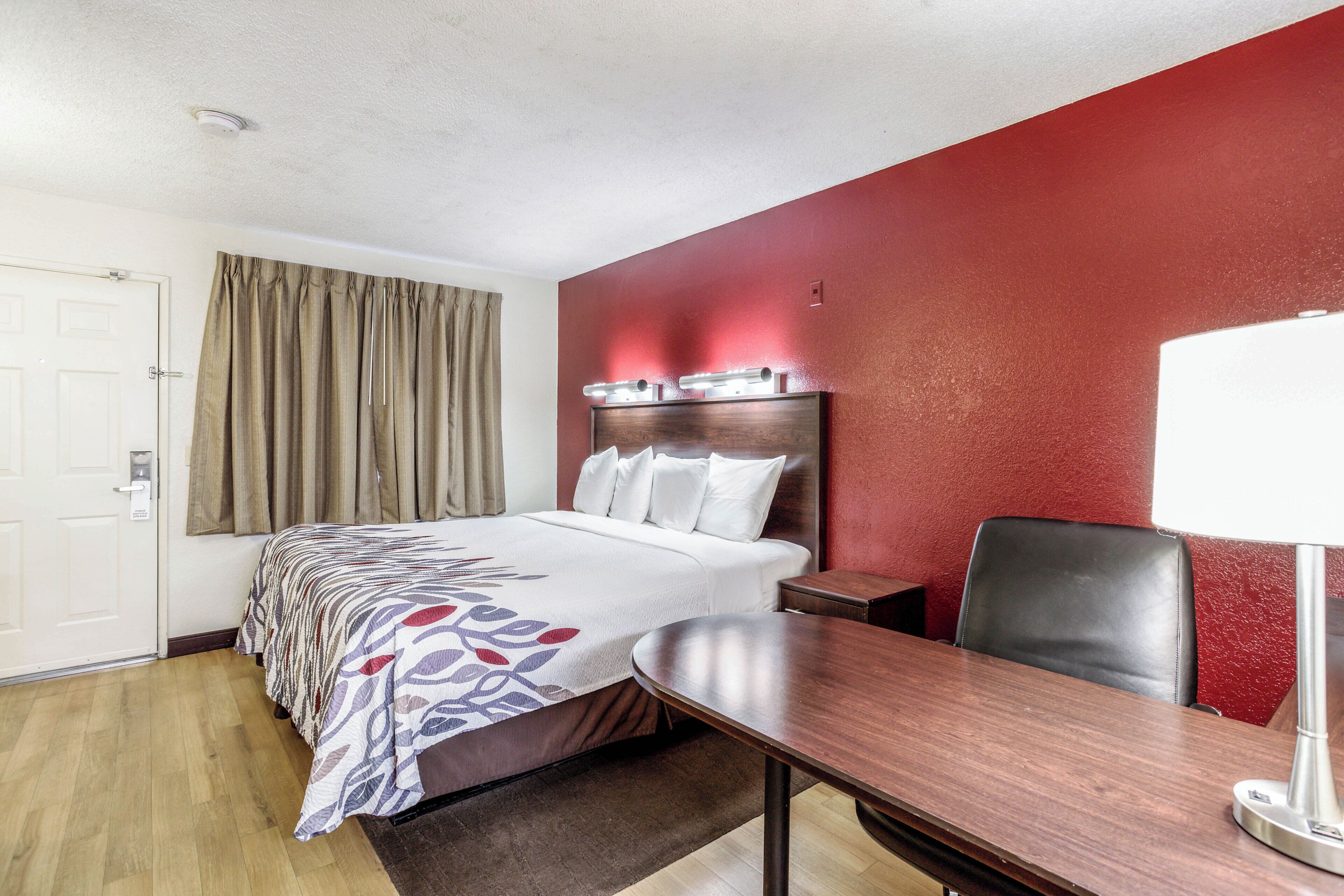 A hotel room with a bed, a desk, a chair, a lamp, and a red accent wall.