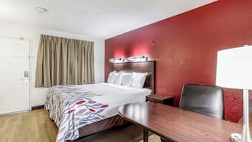 A hotel room with a bed, a desk, a chair, a lamp, and a red accent wall.