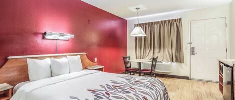 Habitación de hotel con cama, mesita de noche, lámpara, silla, mesa y una puerta.