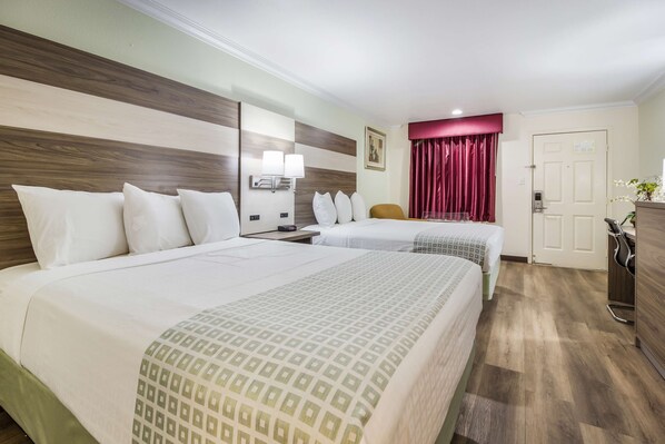 A hotel room with two beds, a wooden headboard, a nightstand with a lamp, a desk with a chair, a door, and a window with curtains.
