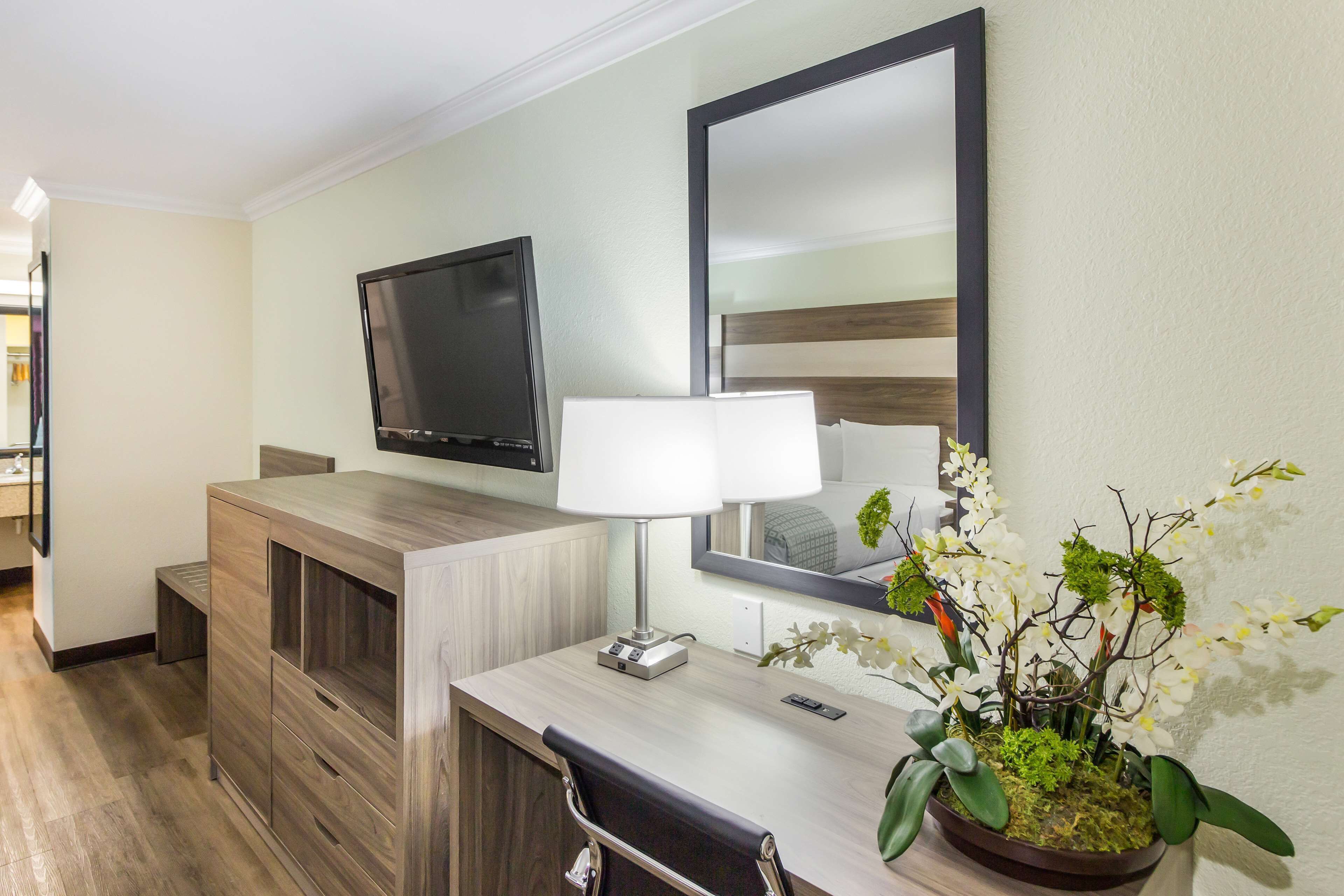Ein Hotelzimmer mit einem Holztisch, einem Flachbildfernseher, einer Nachttischlampe, einem Spiegel und einer Zimmerpflanze mit Orchideen.