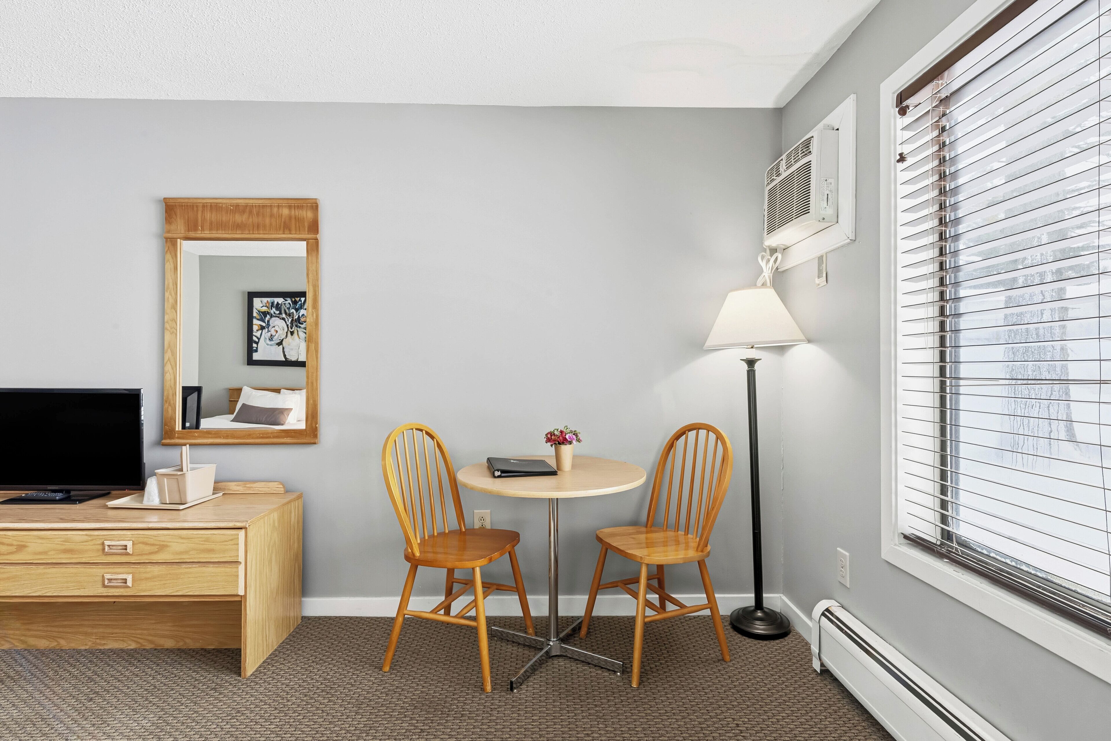 A hotel room with a desk, two chairs, a lamp, a TV, a mirror, and a window with blinds.