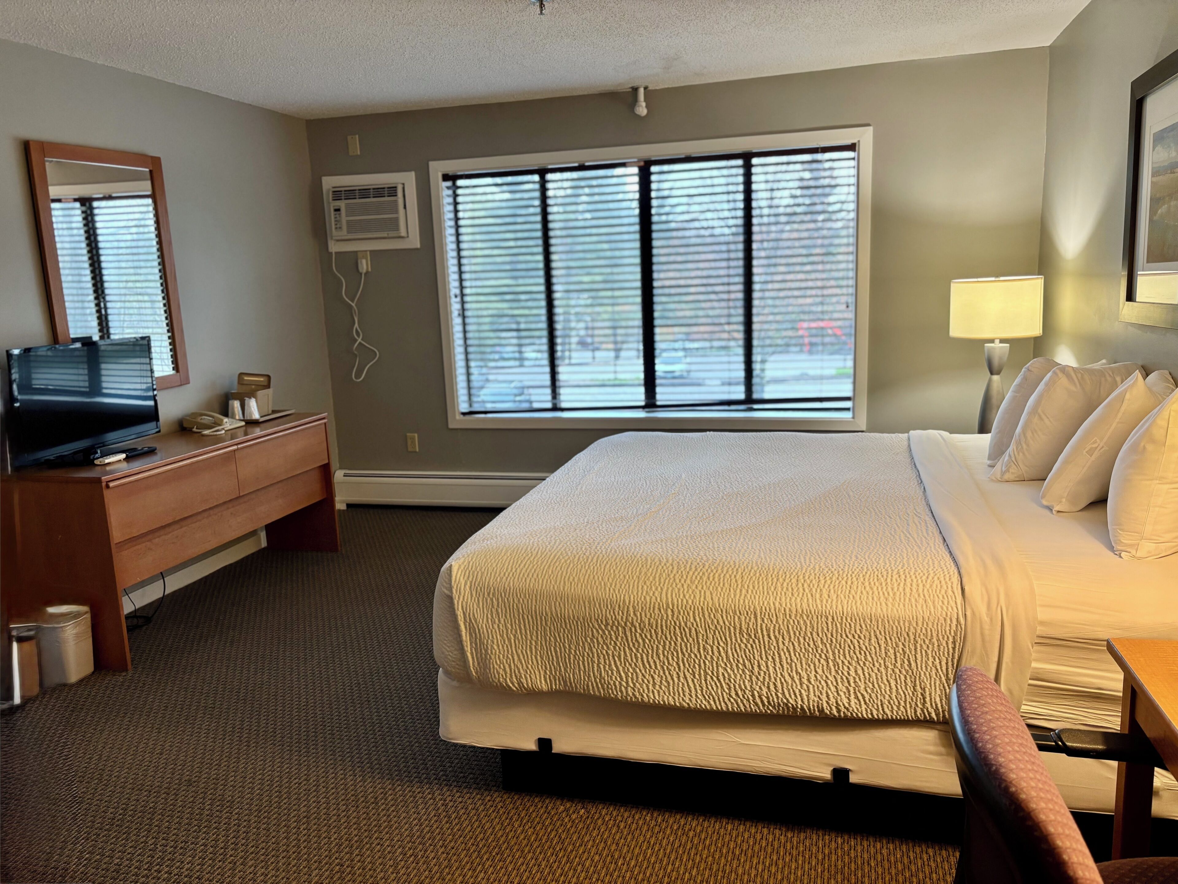 Habitación de hotel con cama, escritorio, televisor, espejo y ventana con persianas.