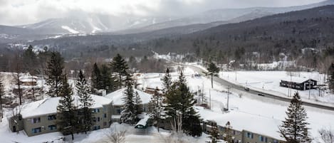 A snow-covered parking lot with several cars, a building with multiple windows, and a mountainous backdrop.