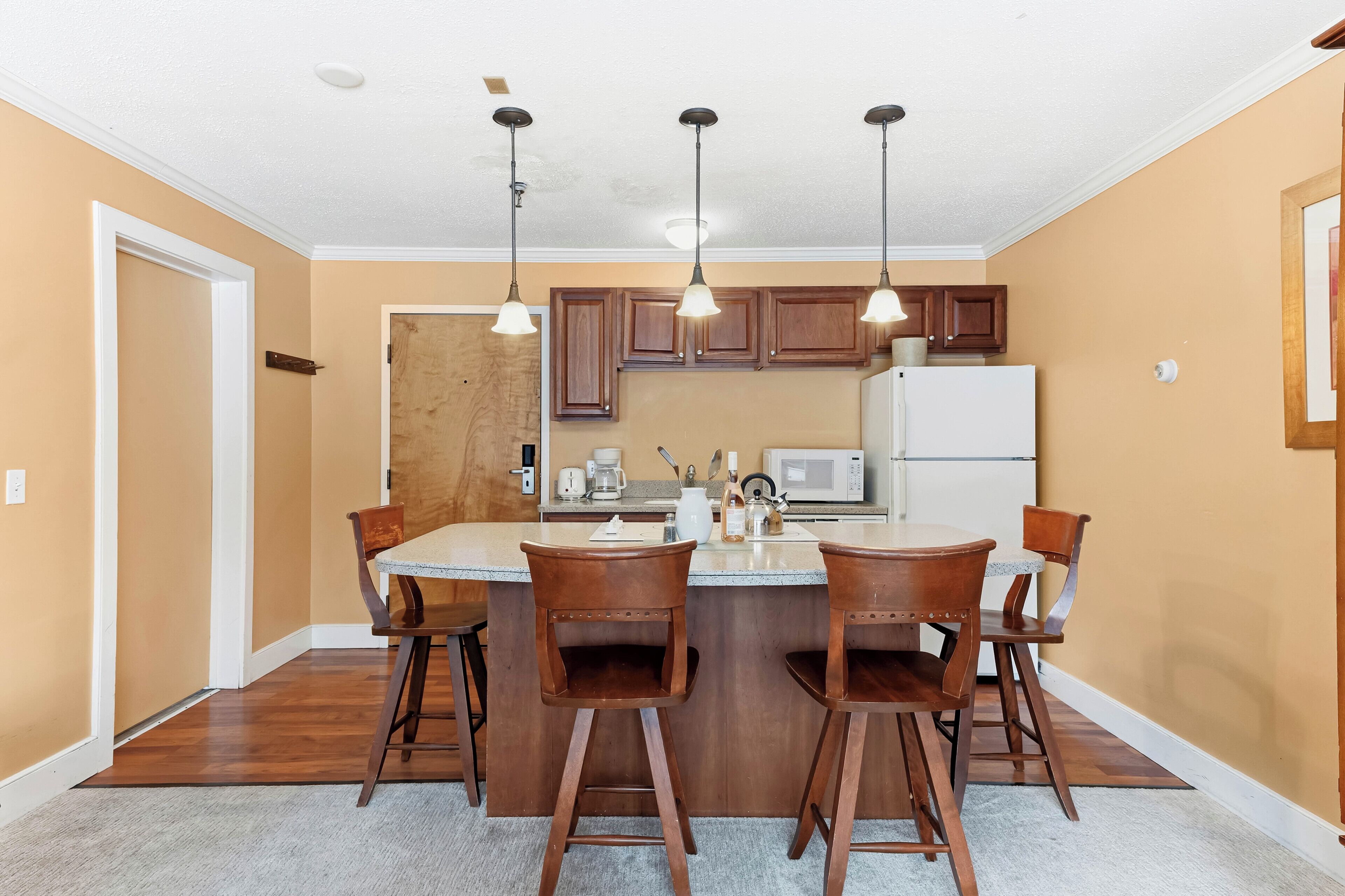 A kitchen with wooden cabinets, a central island with bar stools, pendant lights, a refrigerator, and a microwave.