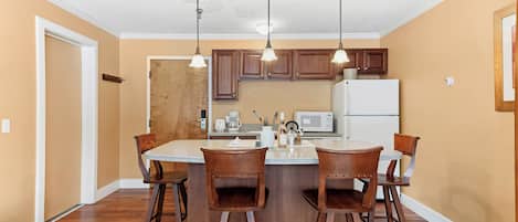 A kitchen with wooden cabinets, a central island with bar stools, pendant lights, a refrigerator, and a microwave.