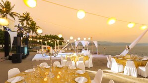 An outdoor wedding venue with round tables set for a reception, string lights overhead, and a view of the ocean.