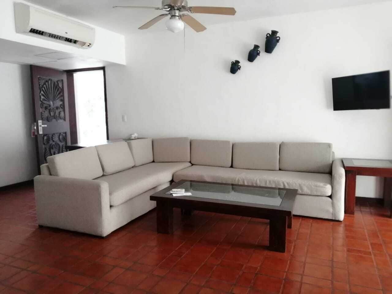A living room with a beige sectional sofa, a glass coffee table, a ceiling fan, and a flat-screen TV mounted on the wall.