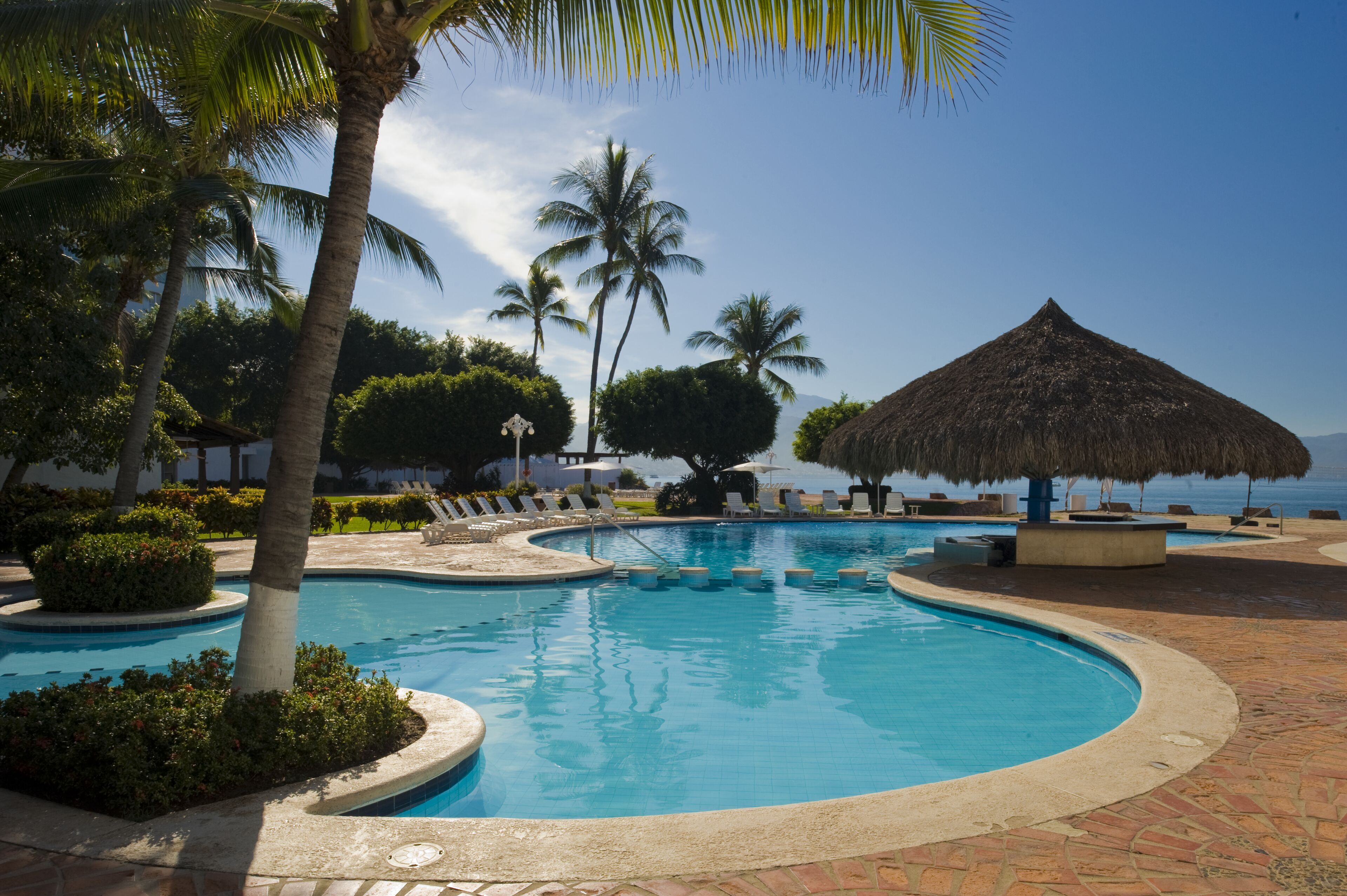 Una piscina con techo de paja, rodeada de palmeras y sillas de descanso, con vista al mar.