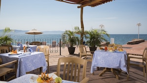 An outdoor dining area with tables and chairs, overlooking a body of water and mountains.