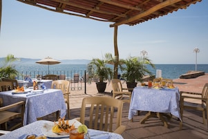 Un área de comedor al aire libre con mesas y sillas, con vistas a un cuerpo de agua y montañas.