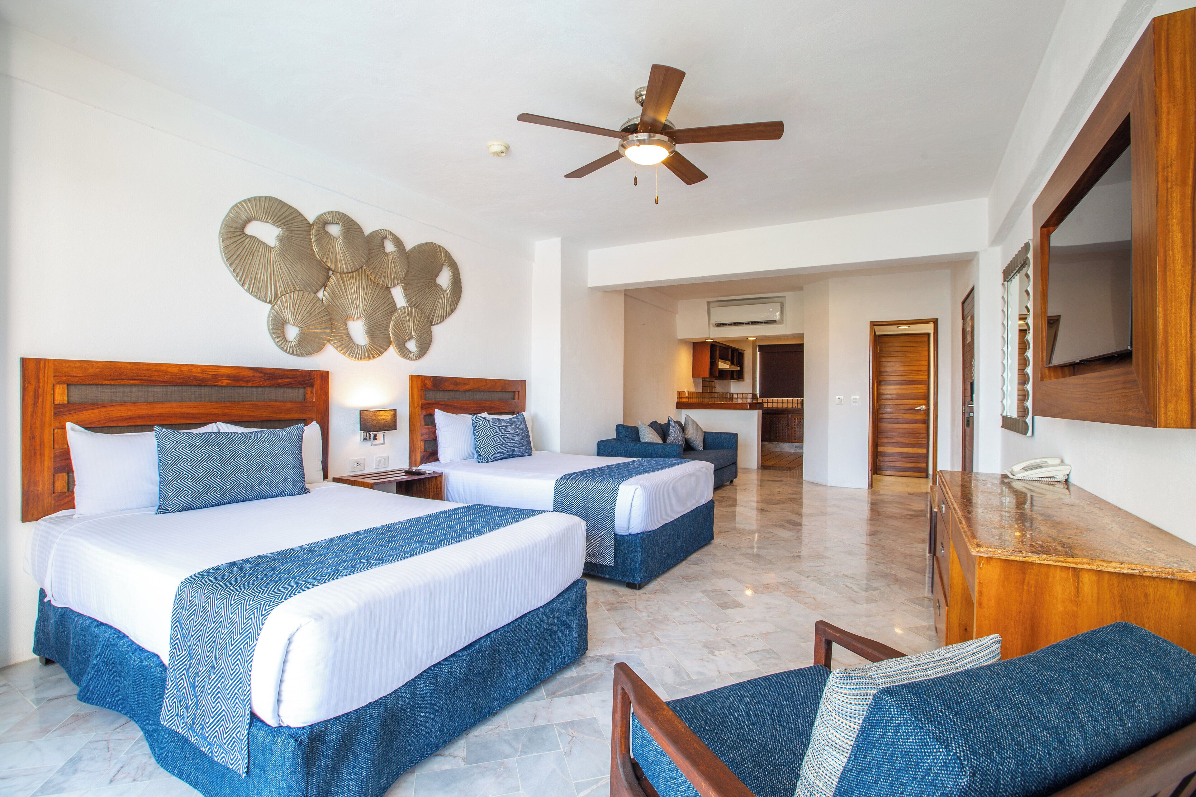 A hotel room with two beds, a ceiling fan, a decorative wall piece, and a view of the kitchen and living area.