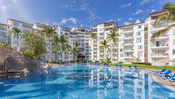 A large swimming pool surrounded by lounge chairs and palm trees, with multi-story buildings in the background.