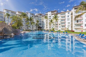 Una gran piscina rodeada de sillas de descanso y palmeras, con edificios de varios pisos al fondo.