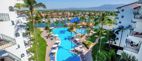 A resort with a large swimming pool surrounded by palm trees, multiple lounge areas, and a view of the surrounding landscape.