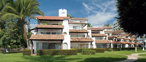 A hotel with multiple white buildings, each with a red-tiled roof and balconies. There are palm trees and well-maintained lawns in the foreground.