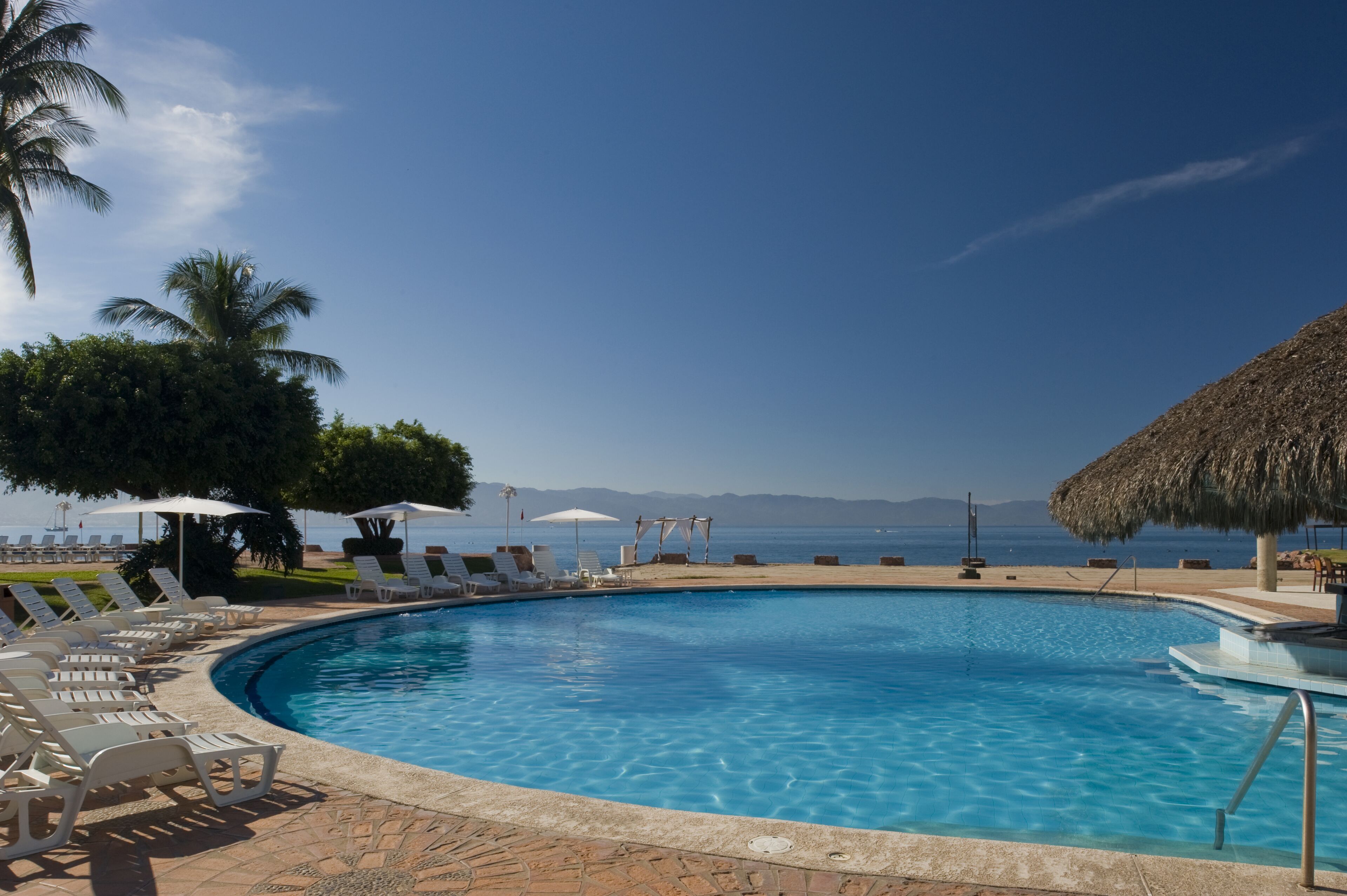 Una piscina azul cristalina con sillas de descanso y sombrillas, una estructura de techo de paja, y una vista al mar y las montañas en el fondo.