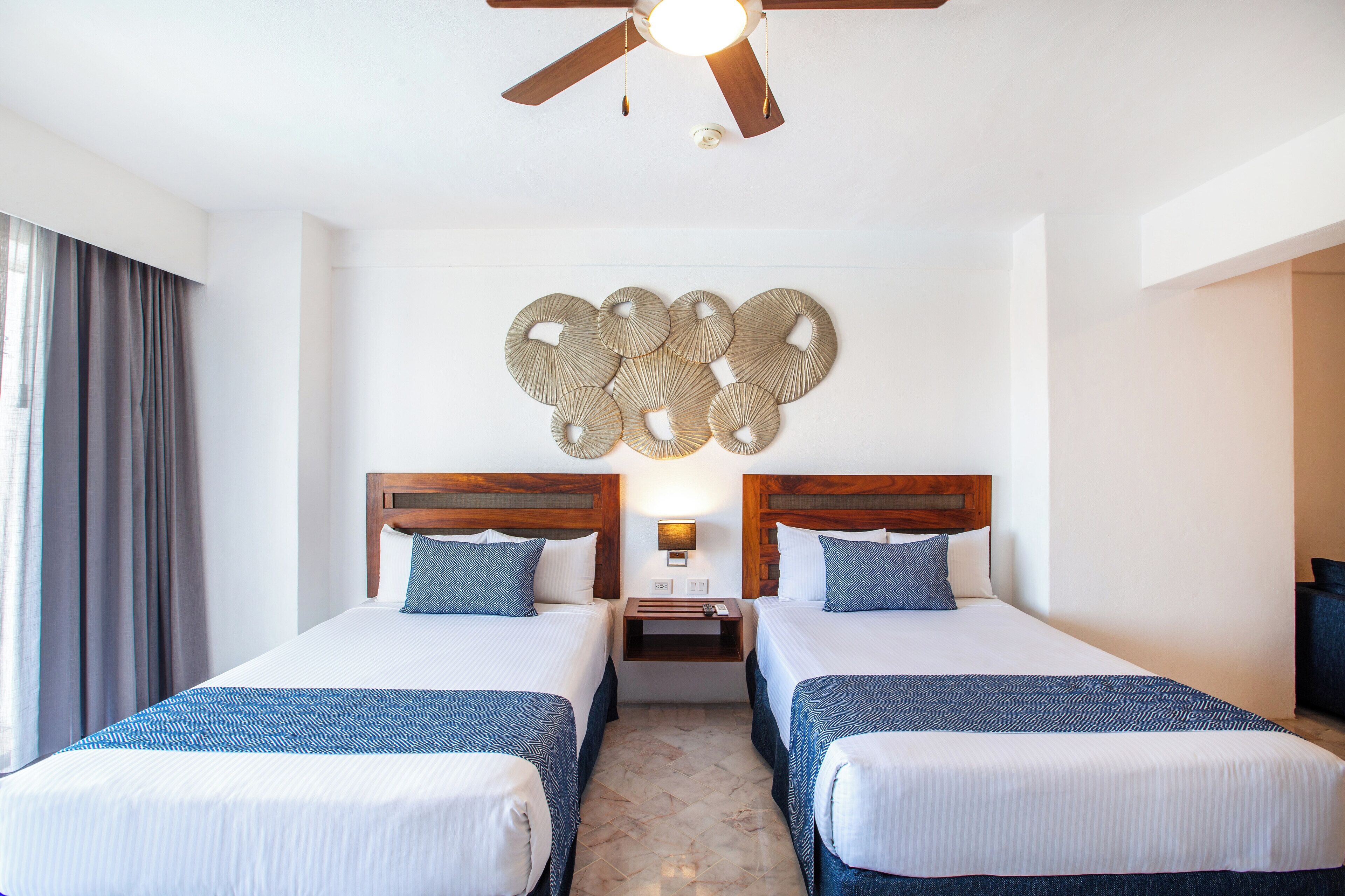 A hotel room with two beds, a ceiling fan, a wall-mounted light, and a decorative wall piece.
