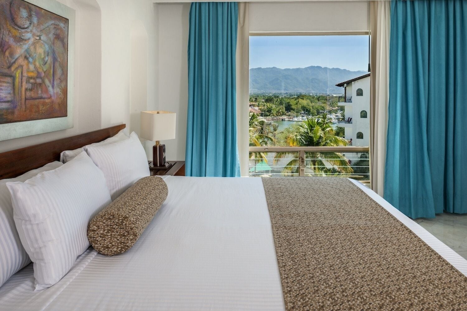 A hotel room with a bed, a painting on the wall, a lamp, and a view of a city and mountains through a balcony.