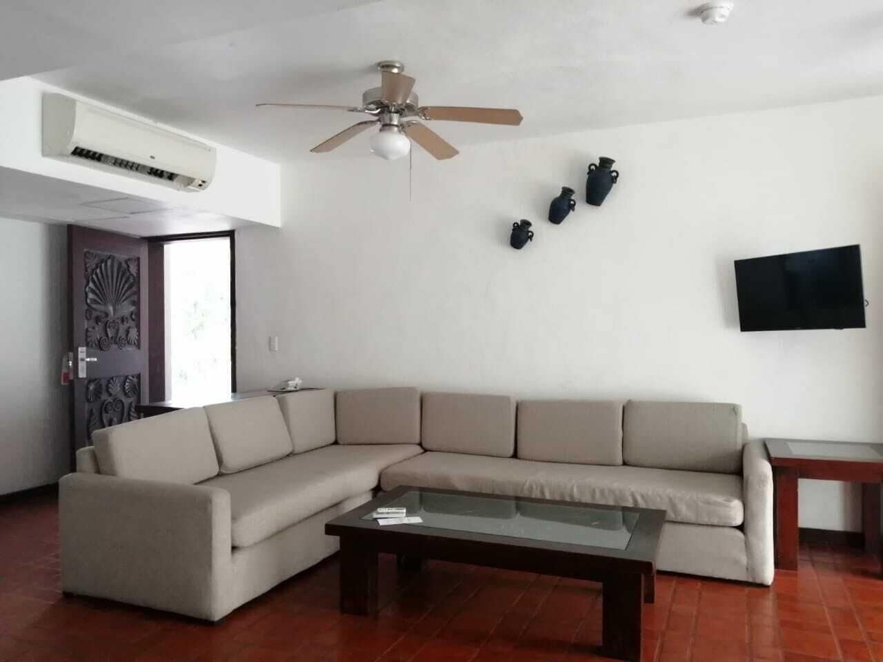 A living room with a beige sectional sofa, a glass coffee table, a ceiling fan, and a flat-screen TV mounted on the wall.