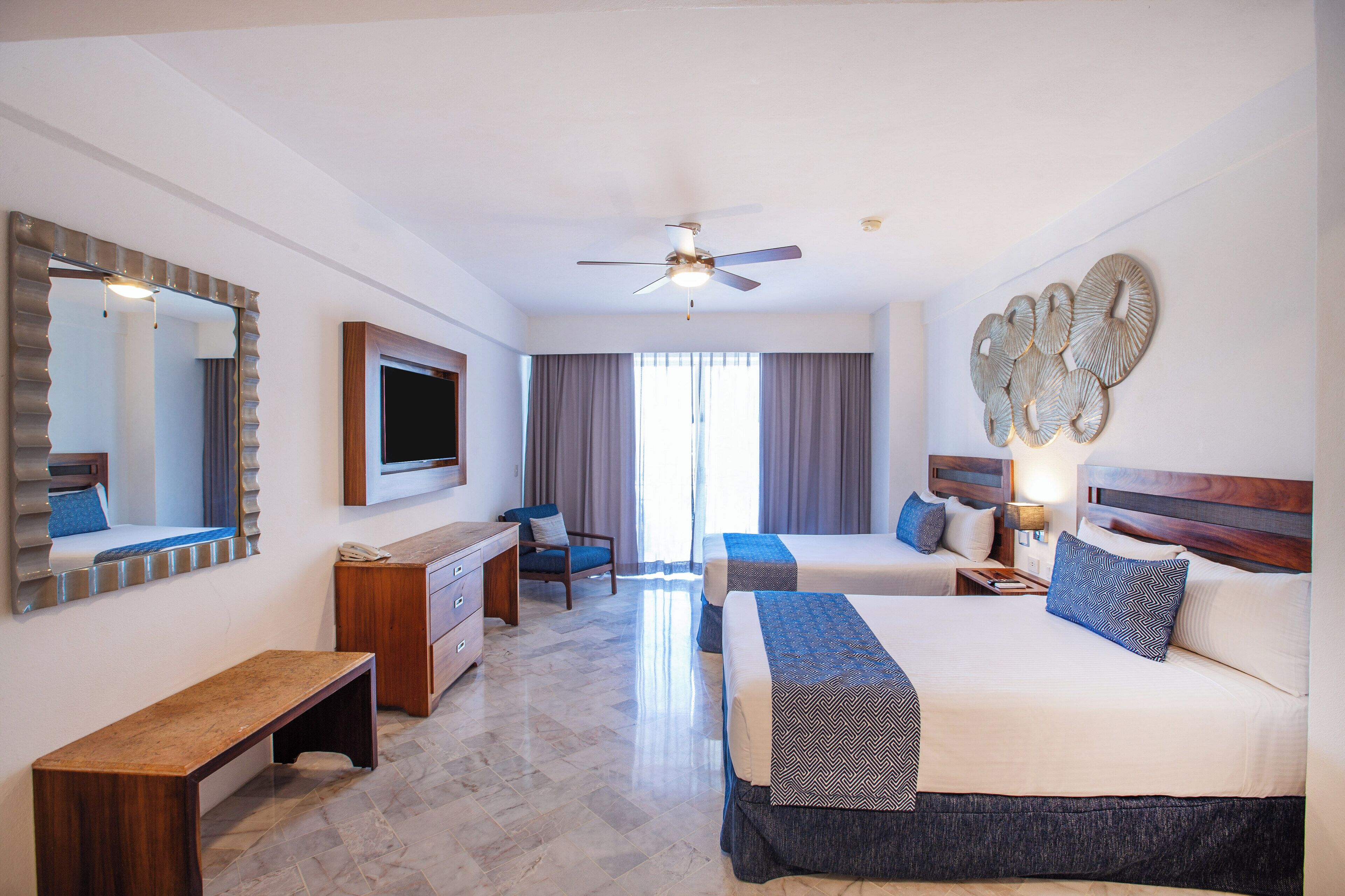 Quarto de hotel com duas camas, uma escrivaninha, uma cadeira, um ventilador de teto e um espelho grande.