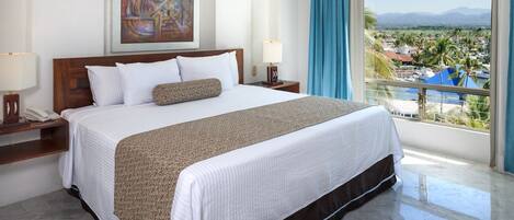 A hotel room with a large bed, a painting on the wall, a view of the city, and a balcony with a view of the mountains.
