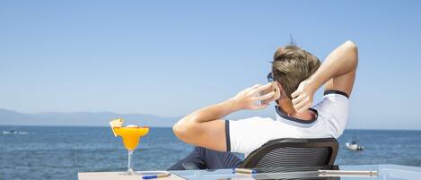 A person sitting at a table by the sea with a drink and a laptop.