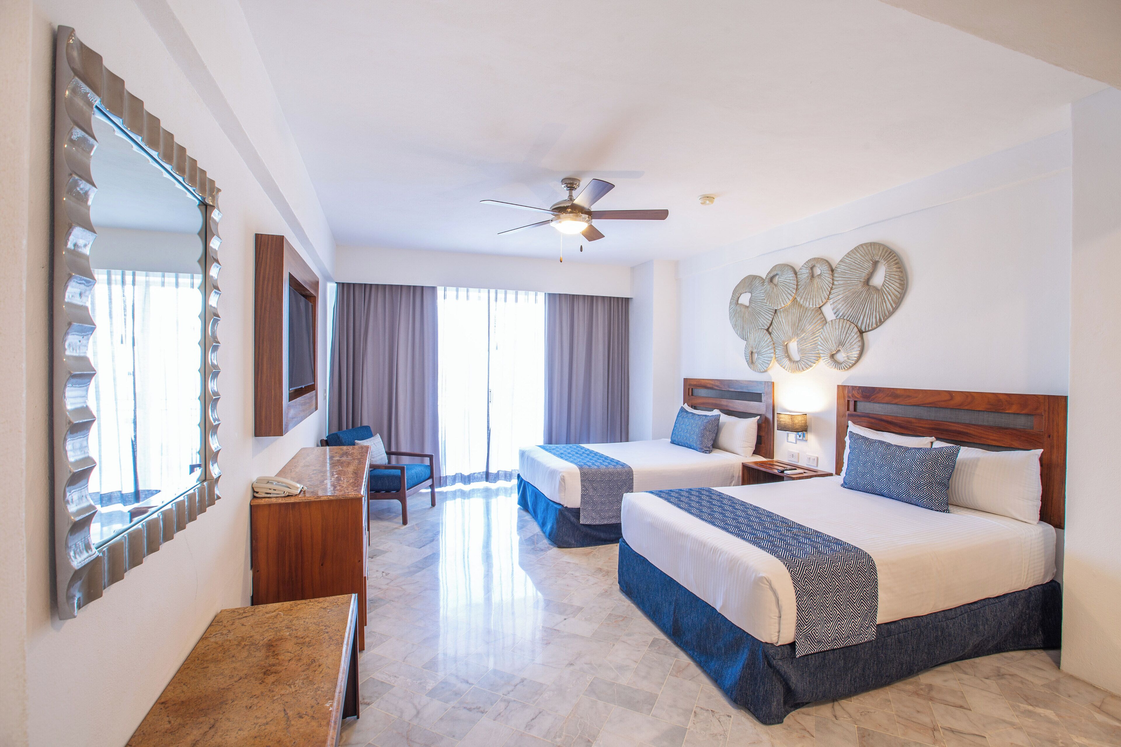 A hotel room with two beds, a ceiling fan, a large mirror, a desk, and a chair.