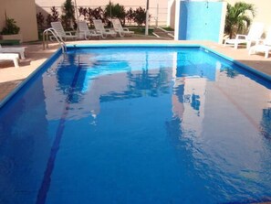 Una piscina rectangular con agua cristalina azul, rodeada de sillas reclinables blancas y un muro de azulejos.