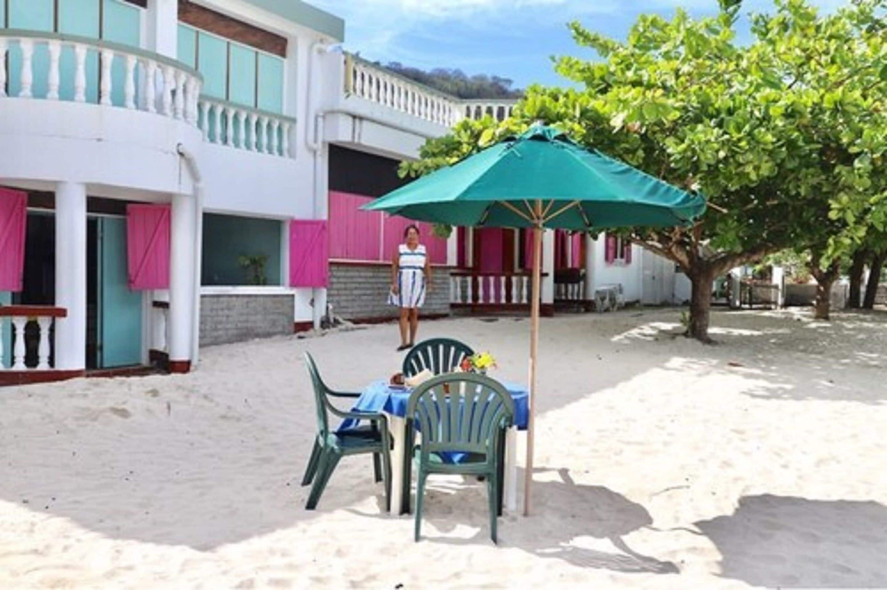 A beachside setting with a table and chairs under a green umbrella, a building with pink shutters, and a tree in the background.