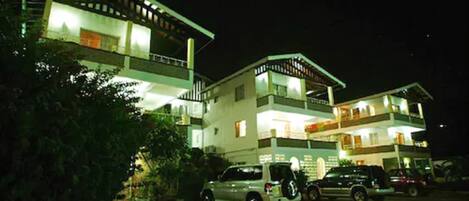 A multi-story building with a flat roof and balconies. There are several cars parked in front.