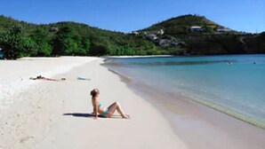 A beach with a person sunbathing, clear blue water, and a hill with greenery in the background.