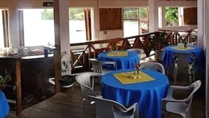 A covered outdoor dining area with tables and chairs, wooden beams on the ceiling, and large windows.