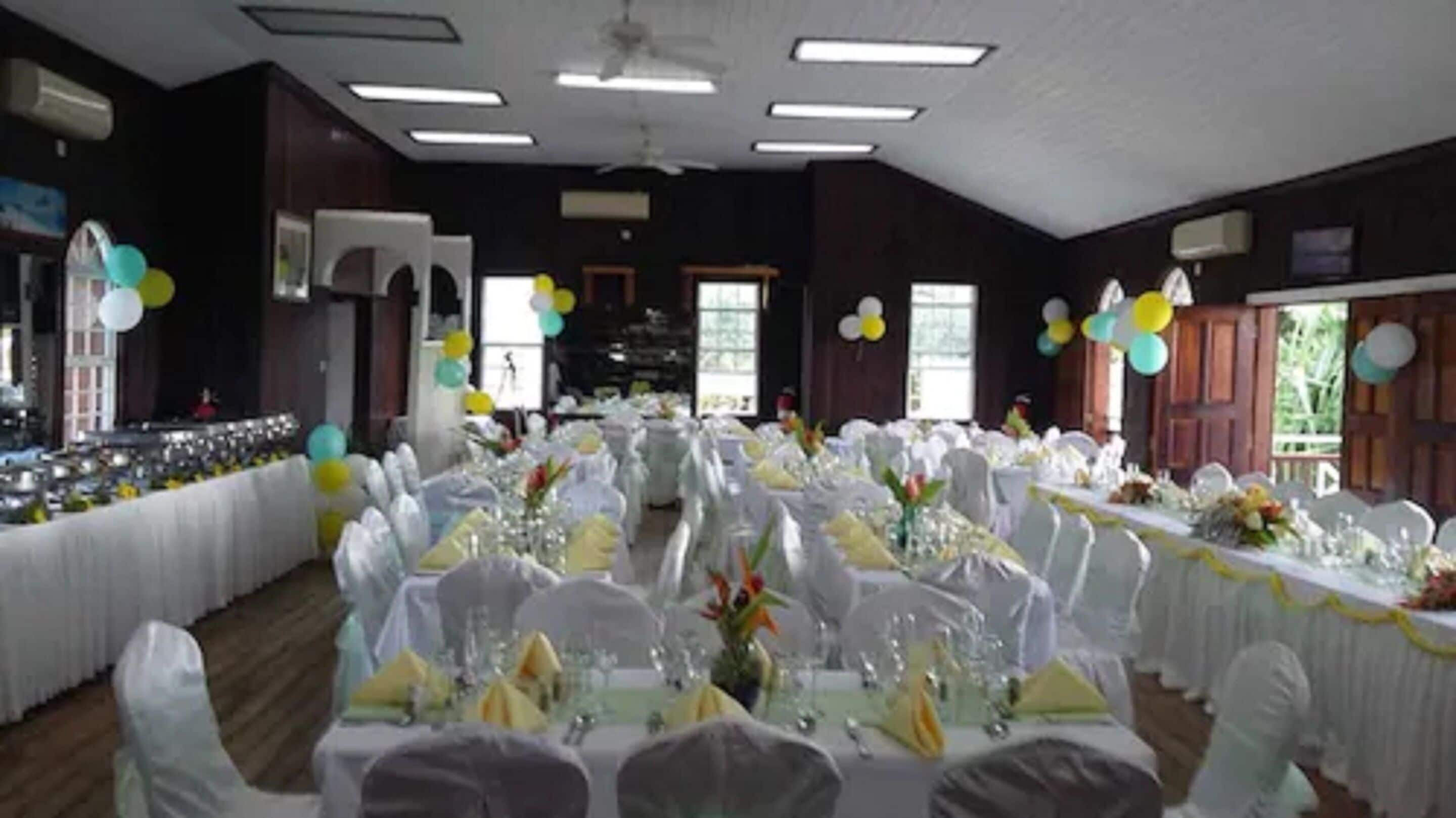 Une salle de banquet où les tables sont dressées pour un événement formel, avec des nappes et des chaises blanches, et des ballons en arrière-plan.