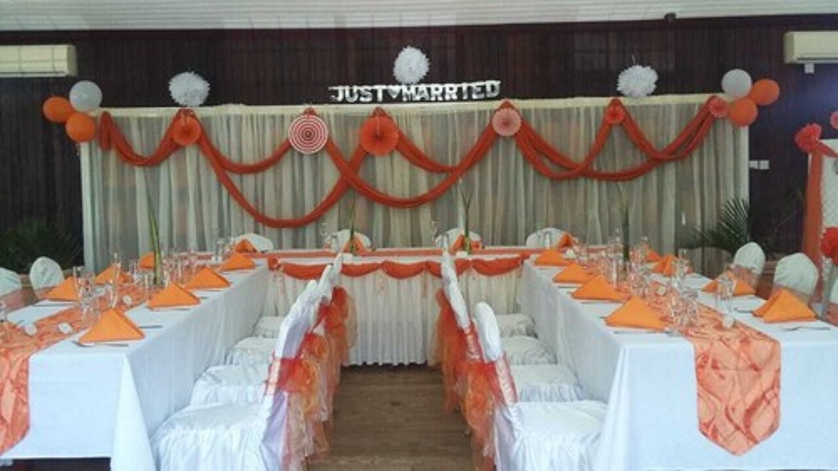 Une salle de réception où les tables sont dressées pour un banquet de mariage, décorée de nappes orange et blanches, et sur laquelle est accrochée une banderole avec l'inscription « JUST MARRIED ».