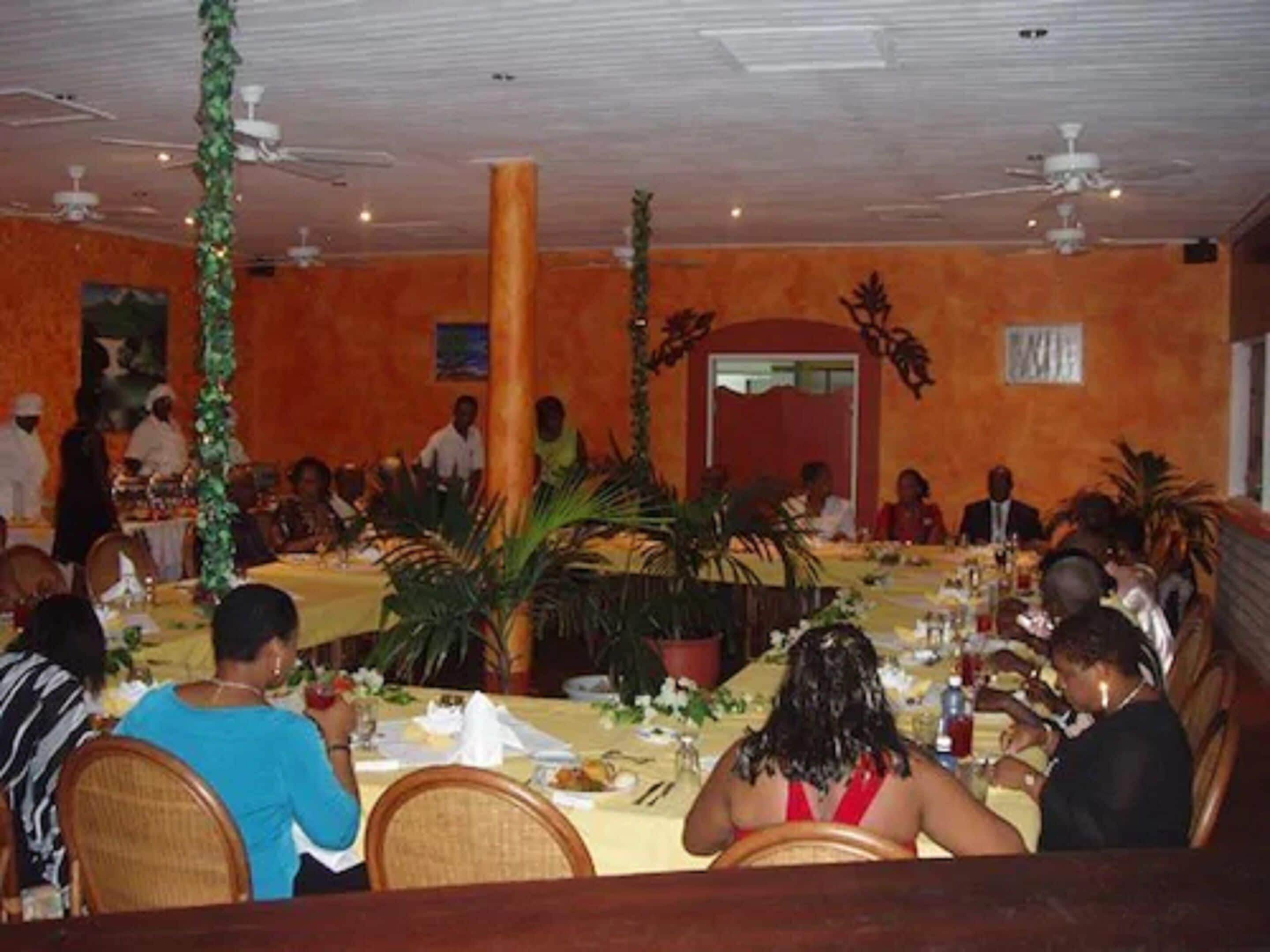 A restaurant with multiple round tables and chairs, decorated with plants and a central pillar.