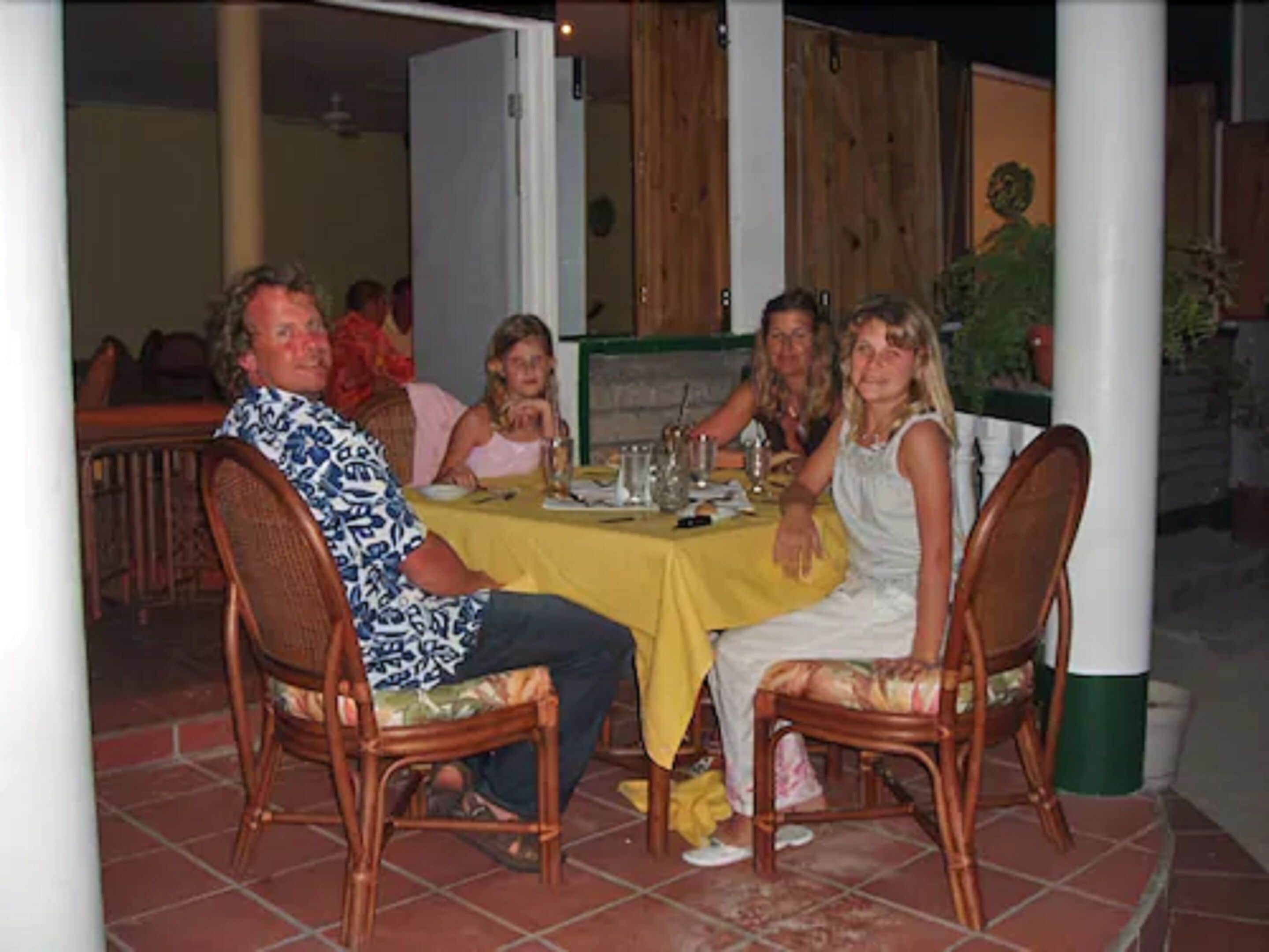 Un grupo de personas sentadas en una mesa al aire libre con mantel amarillo, rodeadas de sillas de mimbre y un suelo de baldosas.